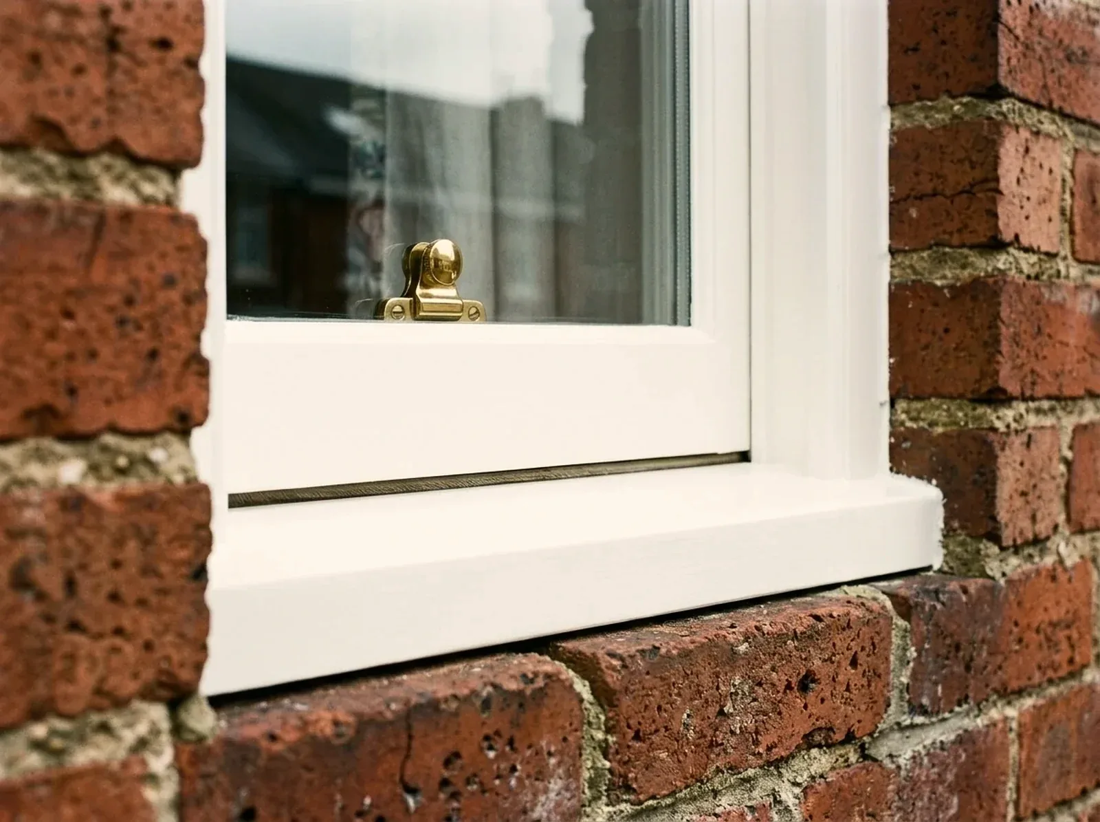 Restored white box-sash in red brick with a brass fitch catch and a brush-pile weatherseal along the bottom rail
